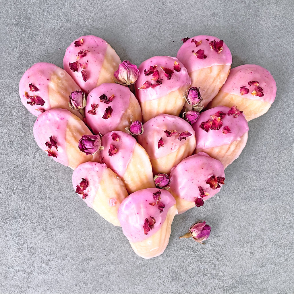 Madeleins mit essbaren Rosenblüten aromatisierter Glasur - in Herzform gelegt auf grauem Hintergrund - nicht nur für Valentinstag - Madeleines mit rosa Glasur - Rezept -